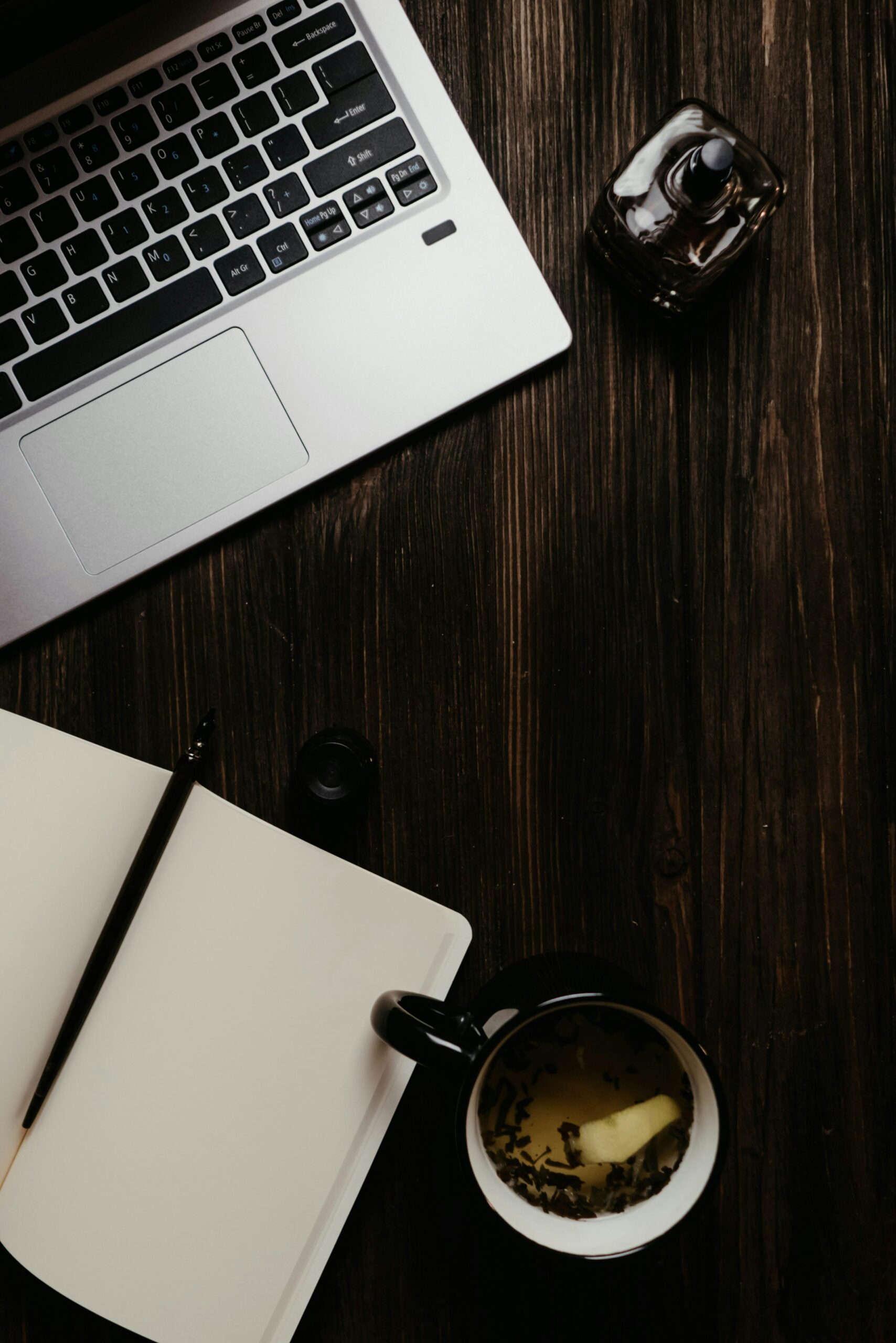 Flat lay of a wooden workspace with a laptop, open notepad, tea, and perfume.